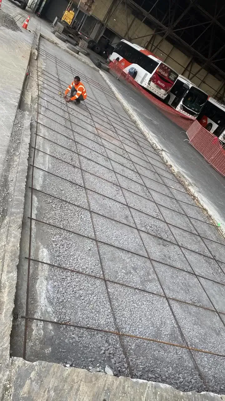 Malla de acero sobre una losa de concreto, con un trabajador y buses al fondo.
