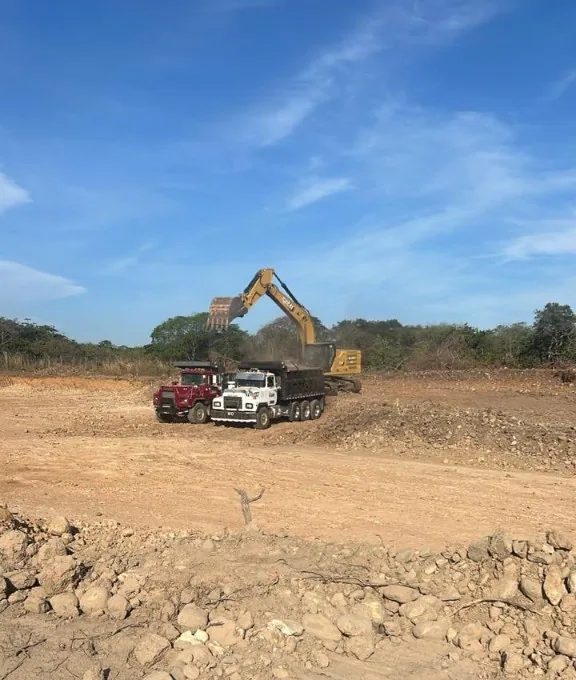 Lote abierto con excavadora y dos volquetes al fondo.