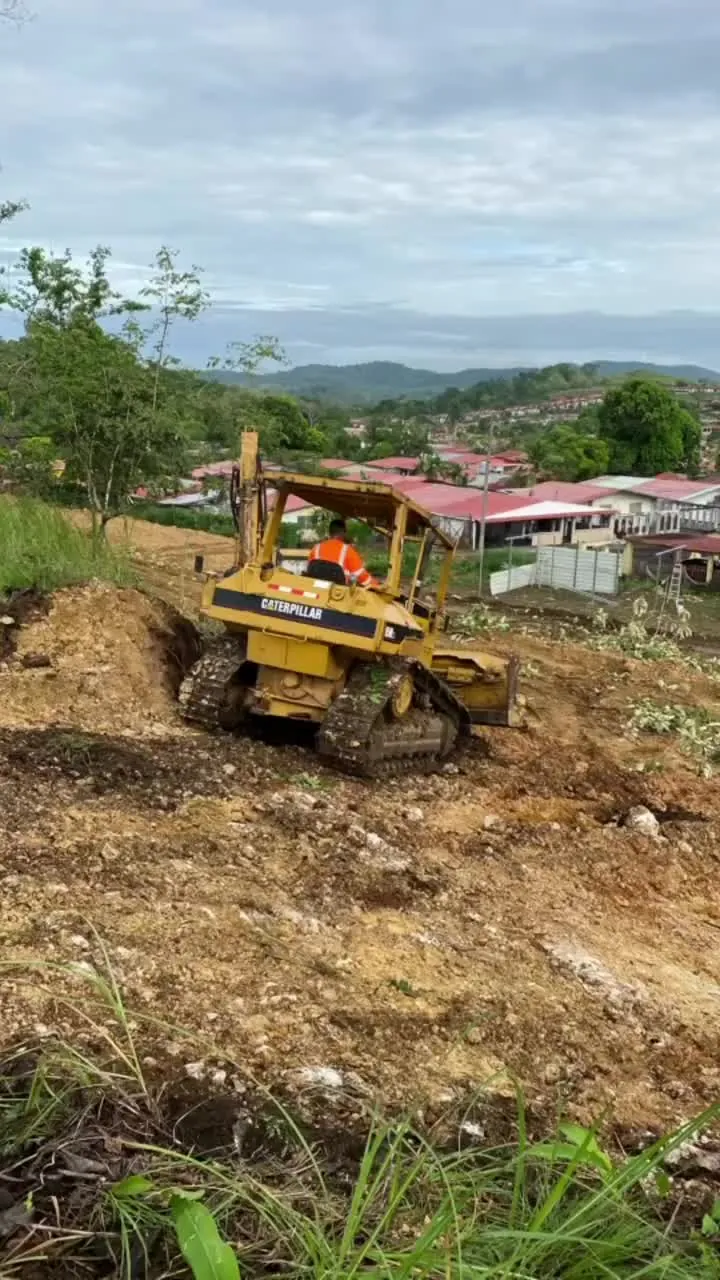 Tractor de orugas Caterpillar empujando tierra, con casas de techo rojo al fondo.