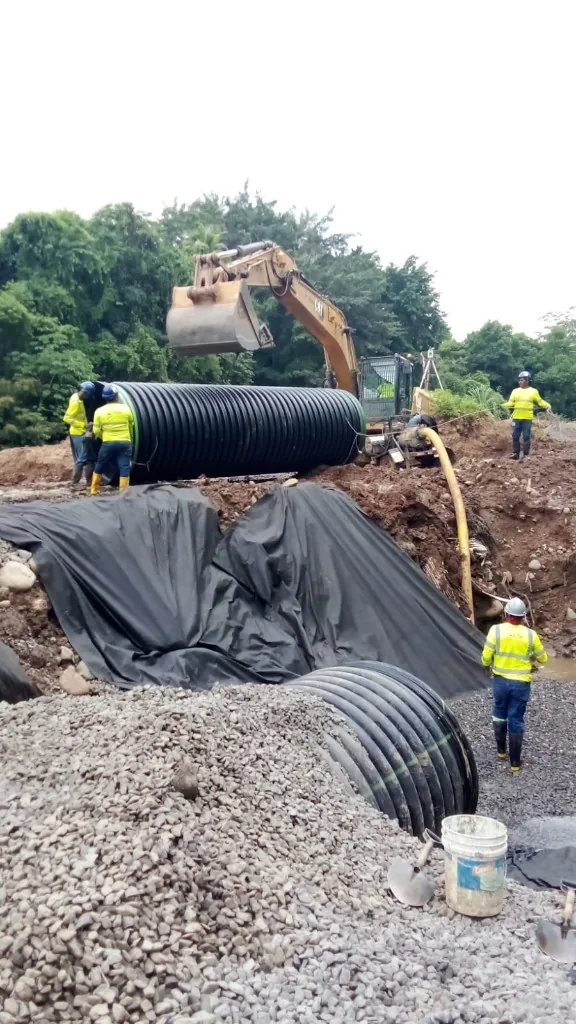 Personal con chaleco colocando un tramo de tubería junto a una excavadora.