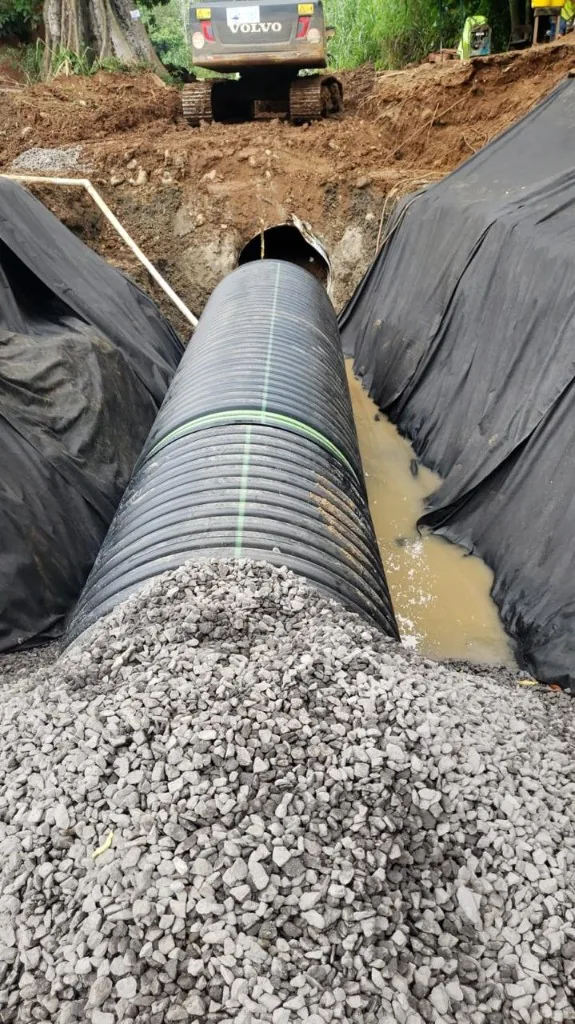Tubería sanitaria tendida en la zanja, con grava al frente y una excavadora atrás.
